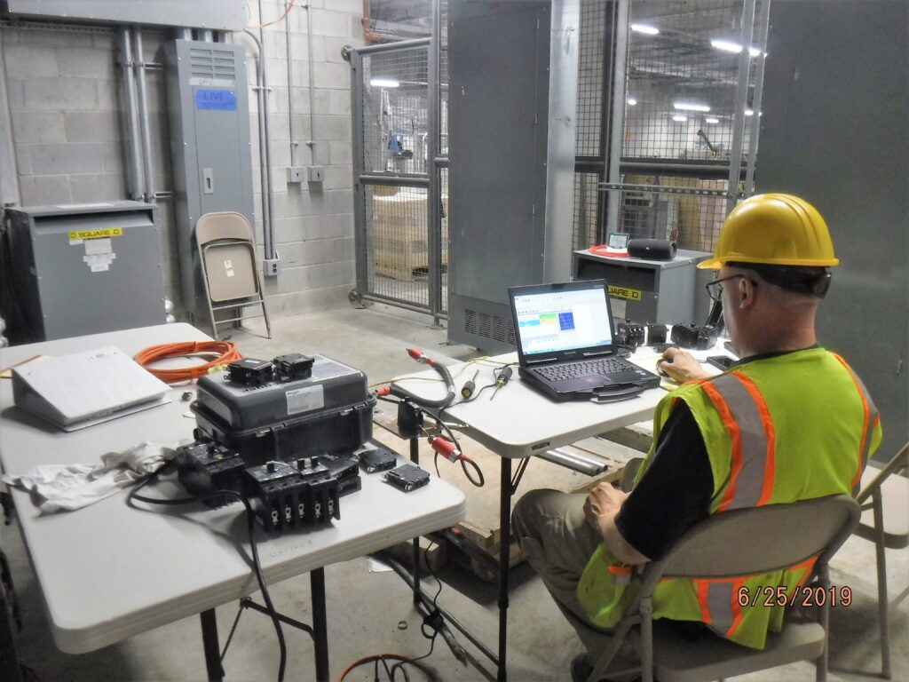 Technician in PPE setting up relay test equipment next to open switchgear