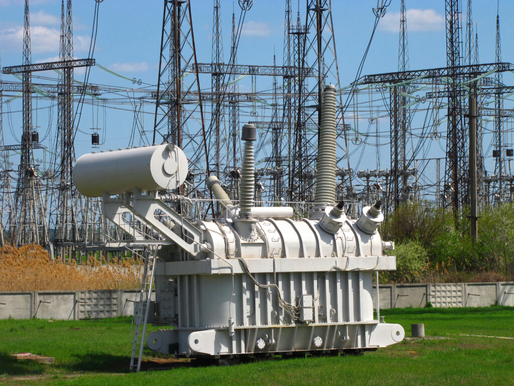 Close-up of a high-voltage power transformer at an electrical substation, with switchyard structures in the background