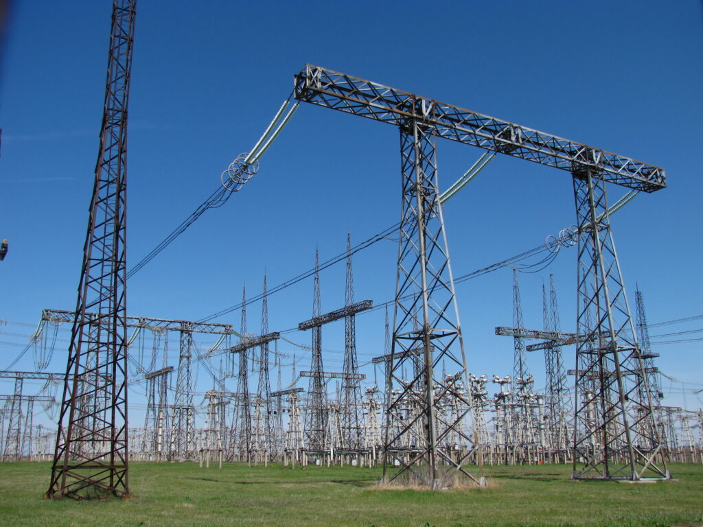 Wide view of an outdoor electrical substation switchyard with transmission towers and high-voltage equipment under a clear sky